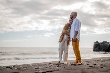 Genç ve sevgi dolu bir çift sahilde ayakta durup sarılıyor. Görüntü, çift ilişkisinin samimiyetini gösteriyor. Görünüşe göre o çift öpüşmek üzere. Yüksek kalite fotoğraf. Bulutlu hava.