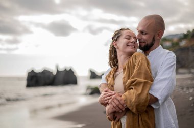 Genç ve sevgi dolu bir çift sahilde ayakta durup sarılıyor. Görüntü, çift ilişkisinin samimiyetini gösteriyor. Görünüşe göre o çift öpüşmek üzere. Yüksek kalite fotoğraf. Bulutlu hava.