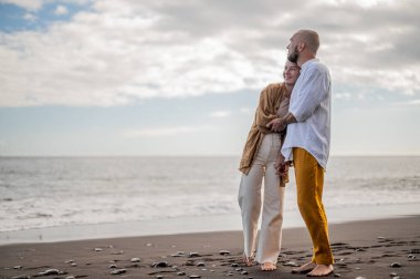 Genç ve sevgi dolu bir çift sahilde ayakta durup sarılıyor. Görüntü, çift ilişkisinin samimiyetini gösteriyor. Görünüşe göre o çift öpüşmek üzere. Yüksek kalite fotoğraf. Bulutlu hava.