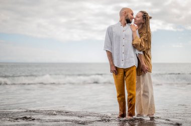 Genç ve sevgi dolu bir çift sahilde ayakta durup sarılıyor. Görüntü, çift ilişkisinin samimiyetini gösteriyor. Görünüşe göre o çift öpüşmek üzere. Yüksek kalite fotoğraf. Bulutlu hava.