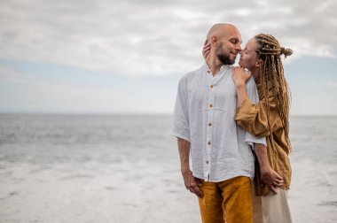 Genç ve sevgi dolu bir çift sahilde ayakta durup sarılıyor. Görüntü, çift ilişkisinin samimiyetini gösteriyor. Görünüşe göre o çift öpüşmek üzere. Yüksek kalite fotoğraf. Bulutlu hava.