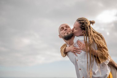 Genç ve sevgi dolu bir çift sahilde ayakta durup sarılıyor. Görüntü, çift ilişkisinin samimiyetini gösteriyor. Görünüşe göre o çift öpüşmek üzere. Yüksek kalite fotoğraf. Bulutlu hava.