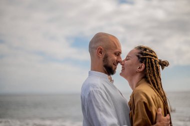 Genç ve sevgi dolu bir çift sahilde ayakta durup sarılıyor. Görüntü, çift ilişkisinin samimiyetini gösteriyor. Görünüşe göre o çift öpüşmek üzere. Yüksek kalite fotoğraf. Bulutlu hava.