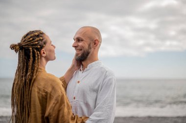 Genç ve sevgi dolu bir çift sahilde ayakta durup sarılıyor. Görüntü, çift ilişkisinin samimiyetini gösteriyor. Görünüşe göre o çift öpüşmek üzere. Yüksek kalite fotoğraf. Bulutlu hava.