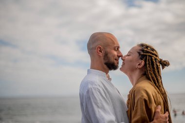 Genç ve sevgi dolu bir çift sahilde ayakta durup sarılıyor. Görüntü, çift ilişkisinin samimiyetini gösteriyor. Görünüşe göre o çift öpüşmek üzere. Yüksek kalite fotoğraf. Bulutlu hava.