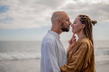 Genç ve sevgi dolu bir çift sahilde ayakta durup sarılıyor. Görüntü, çift ilişkisinin samimiyetini gösteriyor. Görünüşe göre o çift öpüşmek üzere. Yüksek kalite fotoğraf. Bulutlu hava.