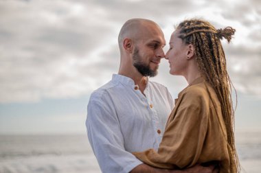 Genç ve sevgi dolu bir çift sahilde ayakta durup sarılıyor. Görüntü, çift ilişkisinin samimiyetini gösteriyor. Görünüşe göre o çift öpüşmek üzere. Yüksek kalite fotoğraf. Bulutlu hava.