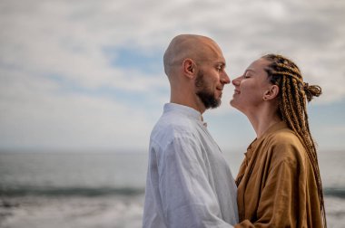 Genç ve sevgi dolu bir çift sahilde ayakta durup sarılıyor. Görüntü, çift ilişkisinin samimiyetini gösteriyor. Görünüşe göre o çift öpüşmek üzere. Yüksek kalite fotoğraf. Bulutlu hava.