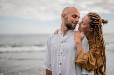 Genç ve sevgi dolu bir çift sahilde ayakta durup sarılıyor. Görüntü, çift ilişkisinin samimiyetini gösteriyor. Görünüşe göre o çift öpüşmek üzere. Yüksek kalite fotoğraf. Bulutlu hava.
