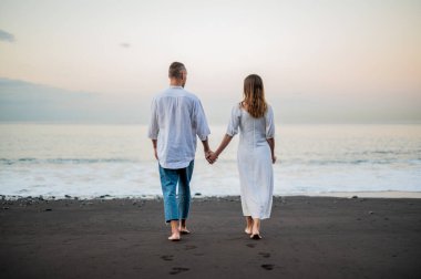 Genç bir çift gün batımında sahilde yürüyor ve el ele tutuşuyorlar, arkadan görüyorlar, beyazlar içinde, tarih ya da yıldönümü