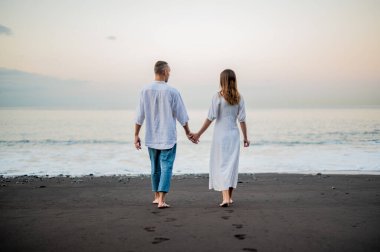 Genç bir çift gün batımında sahilde yürüyor ve el ele tutuşuyorlar, arkadan görüyorlar, beyazlar içinde, tarih ya da yıldönümü