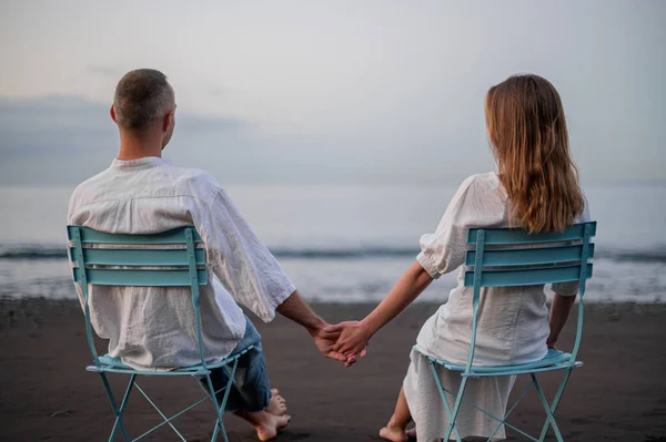 在日落时 年轻夫妇手牵手坐在海滩上 遥望远方 高质量的照片 穿着白色上衣和牛仔裤 — 图库照片