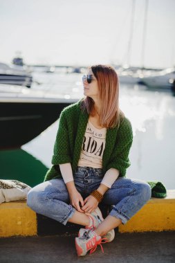 Kot pantolonlu, hırkalı ve güneş gözlüklü genç bir kız deniz kenarında arka plandaki yatlara doğru yürüyor.