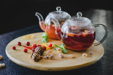 Lokantada siyah bir masanın üzerinde yuvarlak dokulu ahşap bir tahta üzerinde meyveli çay ile iki çaydanlık. Tatlıların, meyvelerin, meyvelerin ve turunçgillerin yanında.