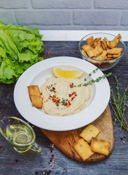 Gri bir masada kesme tahtasında, beyaz bir tabak humus, bir dilim limon, baharatlı baharat ve krakerler marulun yanında, yeşillikler, iki kase, biri zeytin yağlı, diğeri krakerli..