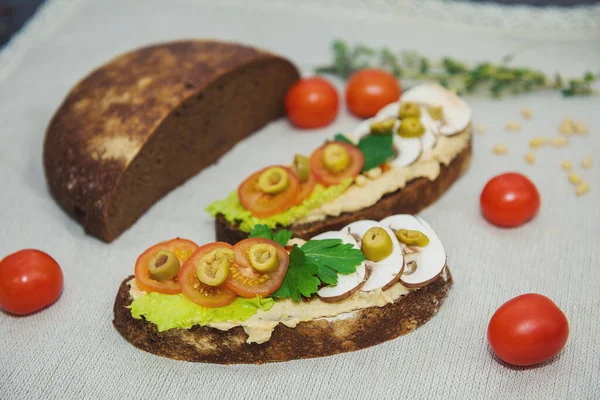 Masanın üzerinde açık bir arka planda yarım somun siyah ekmek, yanında iki sandviç ve humus salatası, domates, zeytin ve bir yumurta var. Ayrıca yakınlarda kiraz domatesleri ve yeşil bir dal var. 