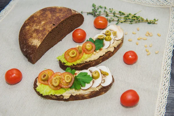 Masanın üzerinde açık bir arka planda yarım somun siyah ekmek, yanında iki sandviç ve humus salatası, domates, zeytin ve bir yumurta var. Ayrıca yakınlarda kiraz domatesleri ve yeşil bir dal var. 