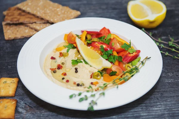 Tahta bir masada gri bir arka planda beyaz bir tabak humus içinde bir dilim limon dilimlenmiş acı biberli domates ve baharatlı baharat. Yakınlarda yarım limonlu ekmek ve yeşil çimen filizleri var.