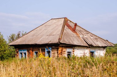 Terk edilmiş bir ev, ormanın yeşilliğinin ve mavi yaz gökyüzünün zeminine karşı otların arasında duruyor.