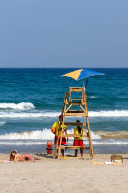 İki banyo güneşli bir yaz günü sahilde güneşlenirken iki cankurtaran kıyıdan izliyor. Bu resimdeki tüm insanlar tanınmaz halde.
