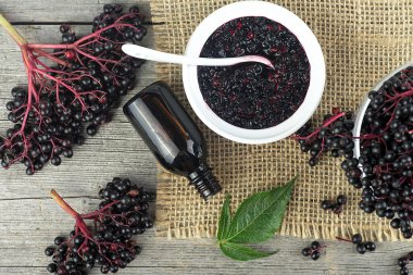 Seramik kavanozda ev yapımı siyah mürver şurubu ve ahşap masa üzerinde yeşil yapraklı siyah mürver demetleri. Mürver şurubu bağışıklığı grip mevsimi için artırılıyor.