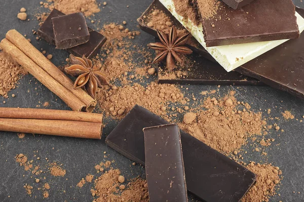 Kırık çikolata parçaları, kakao tozu, tarçın çubukları ve siyah arka planda anason yıldızları.