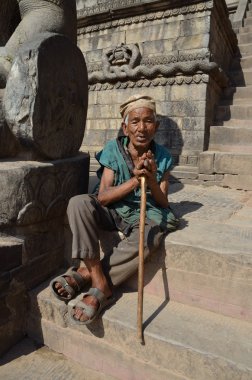 Poor elderly Nepalese man sitting on the steps of the Temple of Taumadhi square in Bhaktapure clipart