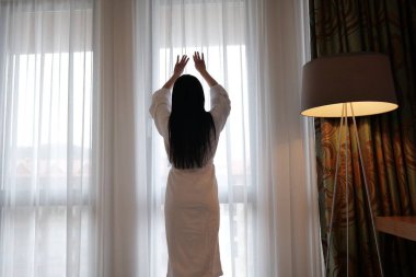 Kadın sabah bir otel odasında perdeleri açar.