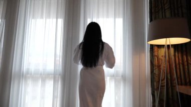 Kadın sabah bir otel odasında perdeleri açar.