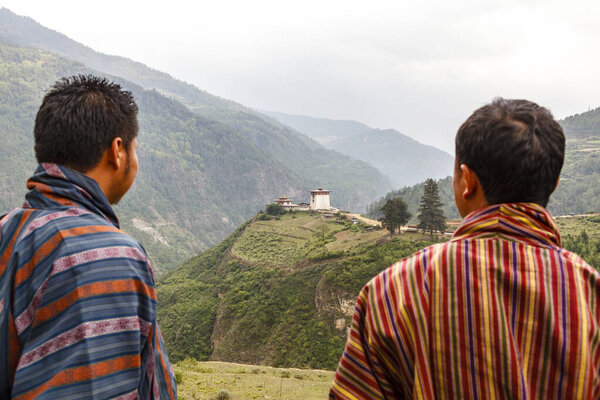 Two men in a gho (Bhutanese traditional clothes)  look at an old monastery in the center of Bhutan, Asia