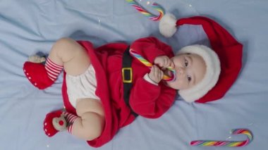 Mutlu Noeller, mutlu yıllar, bebekler, çocukluk, tatil konsepti - yakın plan gülümseyen yeni doğmuş bebek Noel Baba şapkalı, kırmızı beden beyaz yumuşak yatakta oynuyor tatlı Noel şekerini yala.