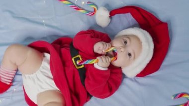 Noel Baba şapkalı, kırmızı elbiseli, beyaz yumuşak yatakta oynayan yeni doğmuş komik bebek tatlı yala. Noel tatili kavramı, şekerler, mutlu yıllar, bebekler, çocukluk. Yakın plan.,