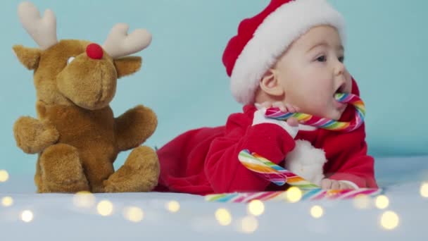 Sorrindo bebê recém-nascido engraçado em chapéu de Papai Noel, jogo de bodysuit vermelho em branco cama macia lamber doce. Férias de conceito Natal, doces, feliz ano novo, bebês, infância. Close-up, — Vídeo de Stock