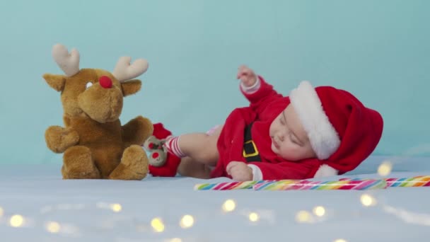 Wesołych Świąt, Szczęśliwego Nowego Roku, Niemowlęta, Dzieciństwo, Koncepcja wakacje - zbliżenie uśmiechnięty śmieszne noworodka w kapeluszu Mikołaja, czerwony body play na białym miękkim łóżku. — Wideo stockowe