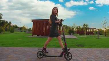 Elbiseli kız elektrikli scooter kullanıyor. Kadın şehir parkında çitli yolda e-scooter ile ilerliyor. Öğrenci işe ya da okula arabayla gidiyor. Kadın bacakları, ekolojik taşıma.