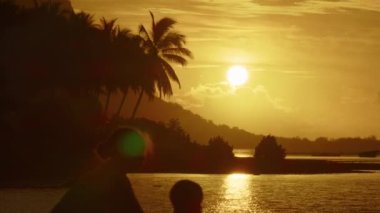 Palmiye ağaçlarının arka planında batan güneşin altında çocukların siluetleri.