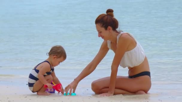 Die junge Mutter und ihre kleine Tochter spielen und entspannen in den Sommerferien am Strand mit Sand. Glückliche Familie, Elternschaft — Stockvideo