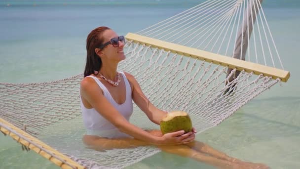 Una muchacha hermosa en un traje de baño blanco relaja mientras que miente en una hamaca y bebe un coco. Playa vacaciones concepto de lujo Océano Índico. Mauricio. — Vídeos de Stock