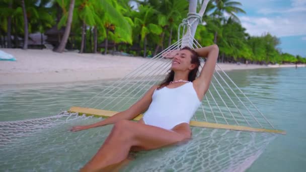 Young female tourist relaxing in a hammock right by the turquoise sea. Caucasian woman on calm summer vacation in Thailand relaxing on the white sand beach by swaying in a rope hammock — Stockvideo