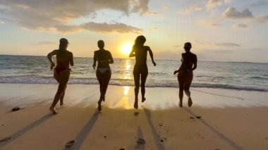 Kumsalda dört kız birlikte koşup zıplıyor, kumsalda mutlu kızlar tatilde.