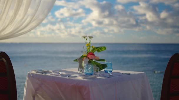 Gün batımında güzel bulutların arkasında sahilde iki kişi için romantik bir akşam yemeği masası. — Stok video