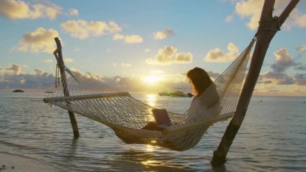 Giovane donna freelance sdraiata su un'amaca con un computer portatile sulla spiaggia tropicale al tramonto sullo sfondo di belle nuvole — Video Stock