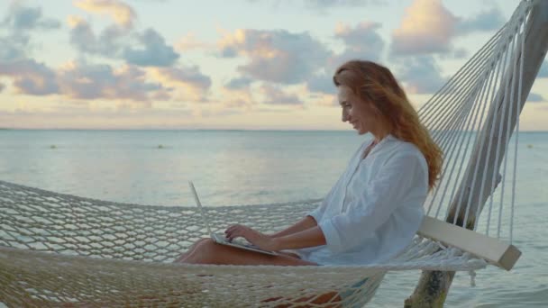 ผู้หญิงสาว freelancer นอนอยู่ในเปลญวนกับแล็ปท็อปบนชายหาดเขตร้อนในเวลาพระอาทิตย์ตกบนพื้นหลังของเมฆที่สวยงาม — วีดีโอสต็อก