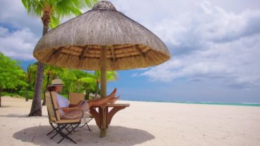 Sarı şapkalı iş kadını, turkuaz suyu olan tropik bir plajın arka planında dizüstü bilgisayar kullanıyor. Tatil, serbest iş, eğlence etkinliği konsepti