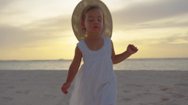 青い海の近くの熱帯のビーチで夏休みの間に白い砂を歩く黄色の帽子の幸せな小さな子供の女の子。笑顔と海でかわいい幼児の子供。背景にある夕日の光 — ストック動画
