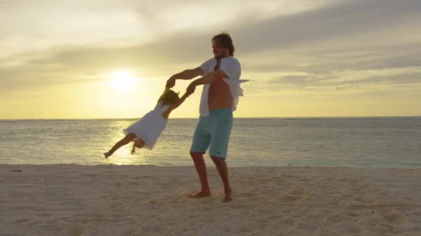 Papà gira sua figlia sulla spiaggia, divertendosi al tramonto. Mio padre gioca con i bambini con l'acqua. Famiglia felice e concetto di infanzia. Silhouette del padre e del bambino sano che sorvola il sole — Video Stock