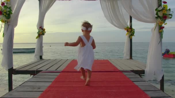 Una niña de dos años con un vestido blanco camina a lo largo de un muelle decorado con flores al atardecer a orillas del Océano Índico. concepto de vídeo de boda — Vídeos de Stock