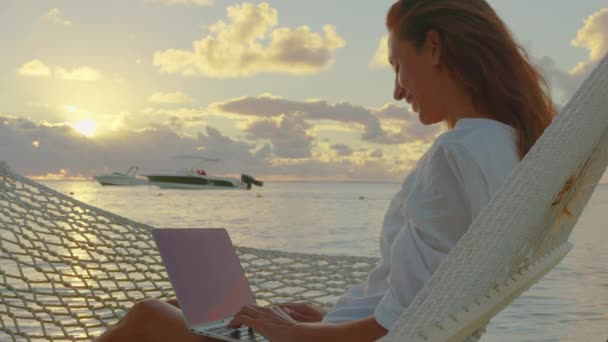 Jonge vrouw freelancer liggend in een hangmat met een laptop op het tropische strand bij zonsondergang op de achtergrond van prachtige wolken — Stockvideo