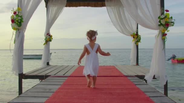 Una niña de dos años con un vestido blanco camina a lo largo de un muelle decorado con flores al atardecer a orillas del Océano Índico. concepto de vídeo de boda — Vídeos de Stock