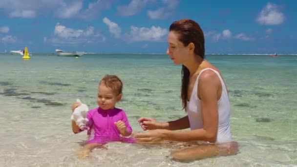 Mamá y su hija están jugando en el agua clara en la orilla del Océano Índico. Mamá y el bebé tienen una gran concha en sus manos. El sonido del océano — Vídeo de stock